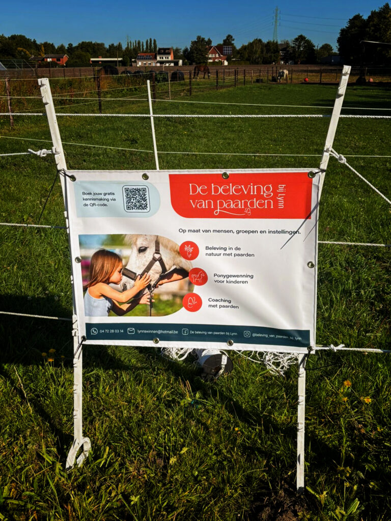 Reclamebanner voor De beleving van paarden bij Lynn, bevestigd aan een omheining in een weide met paarden.