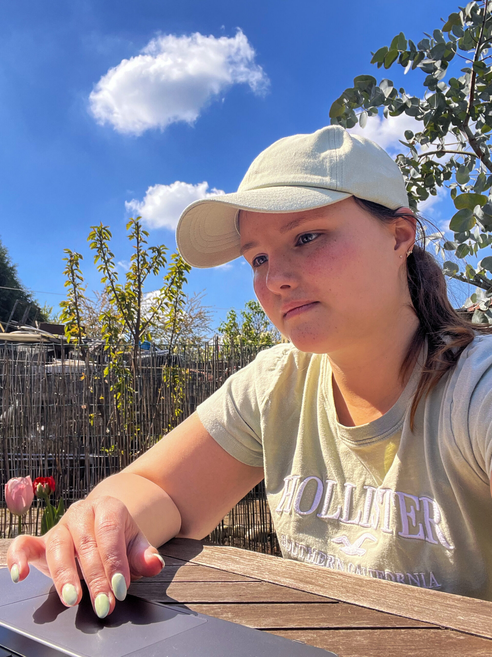 Sarah werkt buiten aan haar laptop, met een tuin en blauwe lucht op de achtergrond.