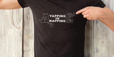 Mockup van een zwart T-shirt met de tekst 'Yapping or napping' en twee kattenillustraties, gedragen door een man.